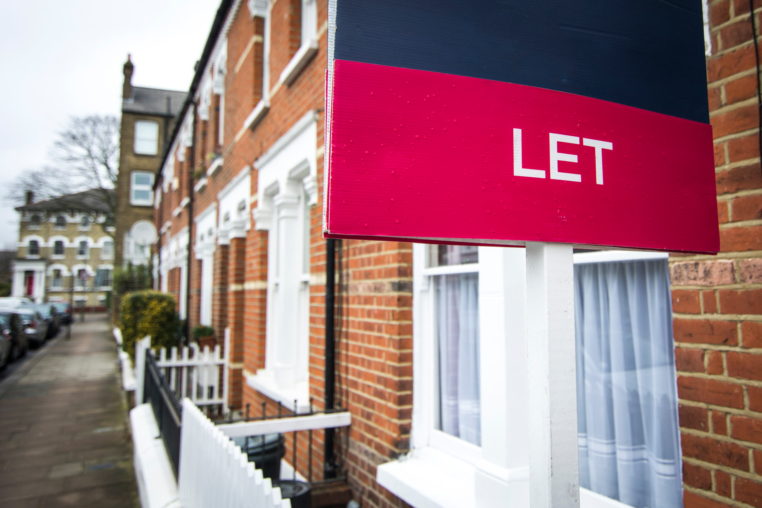 A red and blue sign with the word βLETβ stands in front of a row of brick terraced houses on a residential street.