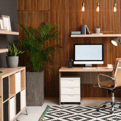 Modern home office with a wooden desk, computer, tan office chair, tall green plants, bookshelves, decorative items, and a patterned rug. Wooden slat accent wall and pendant lights create a cozy atmosphere.