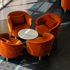 Four orange upholstered armchairs are arranged in a circle around a small round white table on a tiled floor, with a large white planter in the background near a sunlit window.