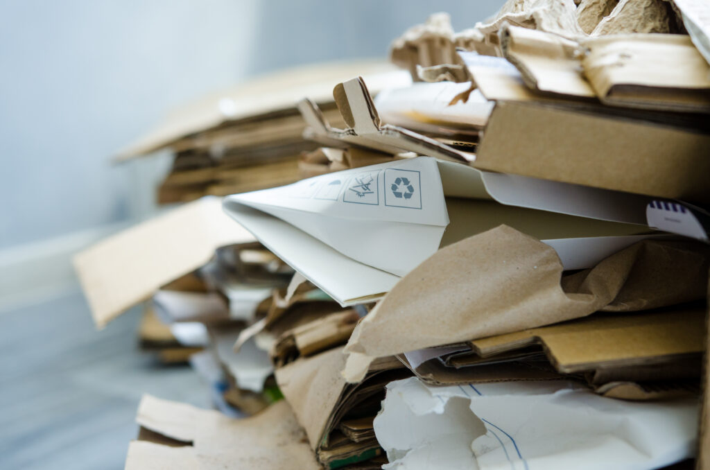 A close-up of a pile of flattened cardboard and paper, some with recycling symbols, stacked for recycling. The background is blurred, focusing on the texture and layers of the materials.