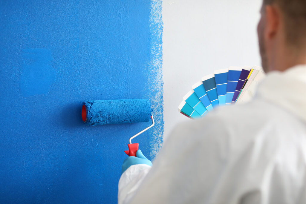 A person in protective clothing paints a wall blue with a roller while holding a fan of blue paint swatches in their other hand.