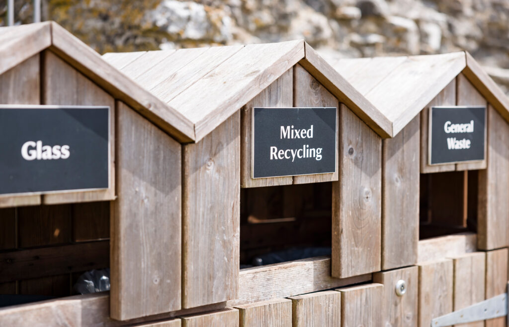 Three wooden recycling bins are labeled "Glass," "Mixed Recycling," and "General Waste," each with a separate slot for disposing of waste, set outdoors on a sunny day.