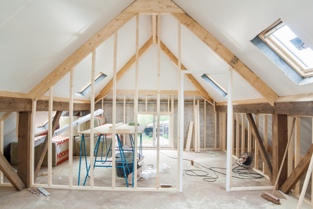 An unfinished attic space with exposed wooden framing and beams, insulation materials, construction tools, and skylight windows letting in natural light. The room is in the process of being renovated.