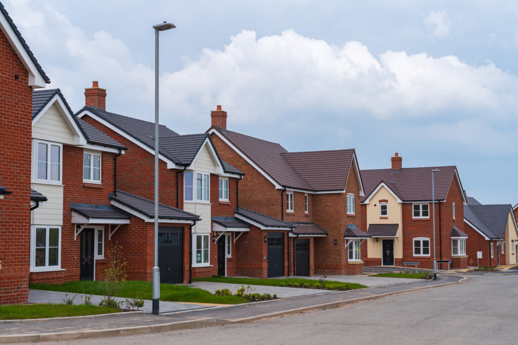 A row of modern suburban houses with brick and cream exteriors, neatly kept front lawns, and driveways, set along a quiet residential street under a partly cloudy sky.