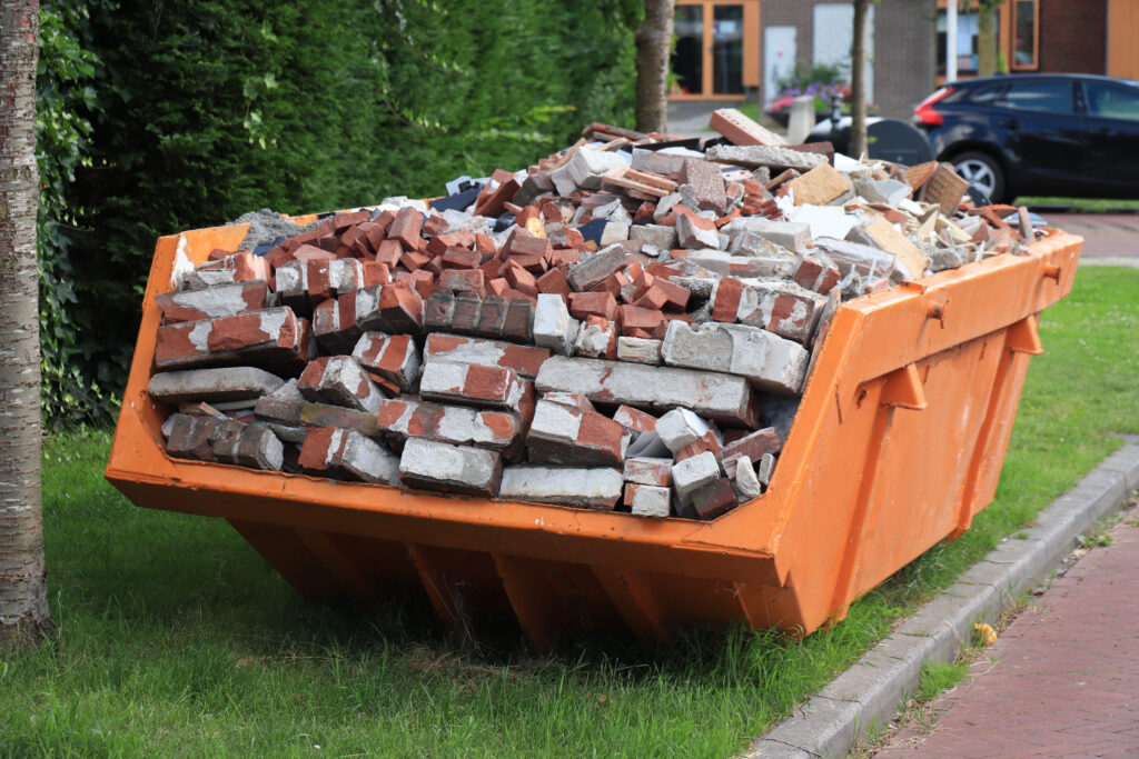 An orange dumpster filled with broken bricks and construction debris sits on grass beside a sidewalk in a residential area.