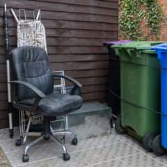A worn black office chair and an ironing board are placed outside next to green and blue wheelie bins, against a brown wooden shed wall. The ground is covered with a patterned mat.