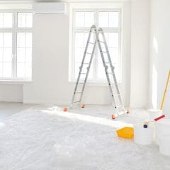 A bright, empty room with large windows, a folded ladder, and painting supplies including buckets, a roller, and plastic sheets covering the floor, indicating ongoing renovation or painting work.