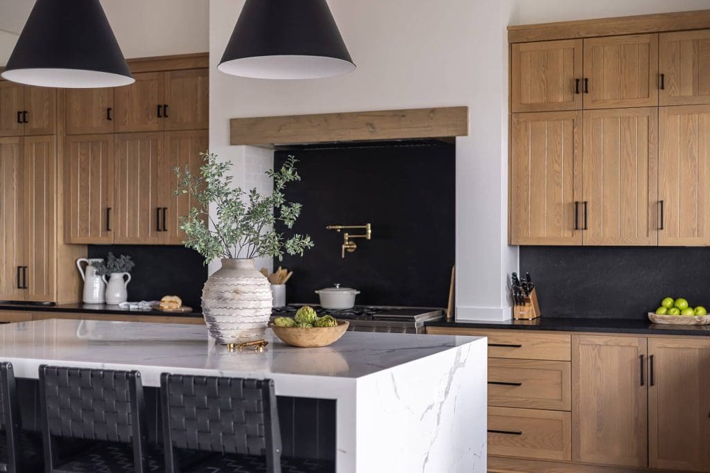 Modern kitchen with light wood cabinets, a white marble island, black barstools, black backsplash, and gold fixtures. A decorative vase with greenery and bowls of fruit are on the island under two black pendant lights.