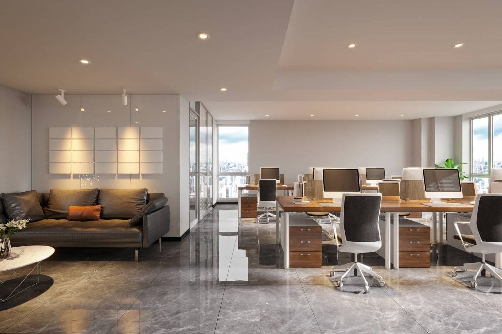 Modern open-plan office with several workstations, black swivel chairs, computers, and wooden desks. A lounge area with a gray sofa and coffee table is on the left. Large windows provide city views and natural light.